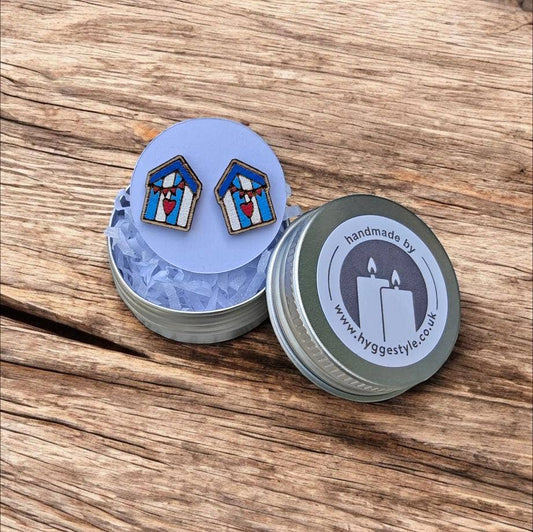 Two small rectangular stud earrings with a blue and white beach hut design and red hearts, displayed on a round light blue backing card above two silver tins on a wooden surface. One tin is open showing the earrings, and the other is closed with a lid label reading 'handmade by www.